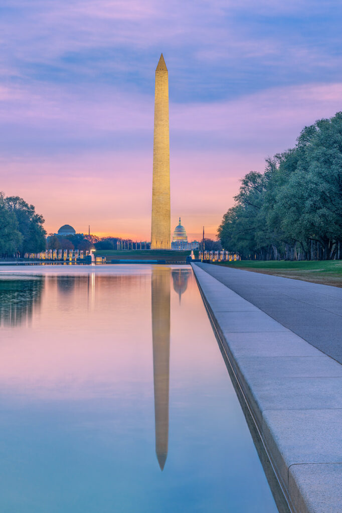 The Washington Monument is lit up by lights at sunset. You can also see the reflection of it in the water and the capital building behind it.