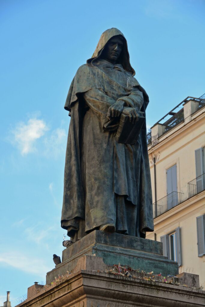 This is a statue of Giordano Bruno. The statue itself is a man wearing a cloak. He is holding a book in one hand and his wrist with the other as he gazes down at the ground. The statue is grey and there is a blue sky with a few white fluffy clowns in the background as well as a pale yellow building with windows and balconies. 