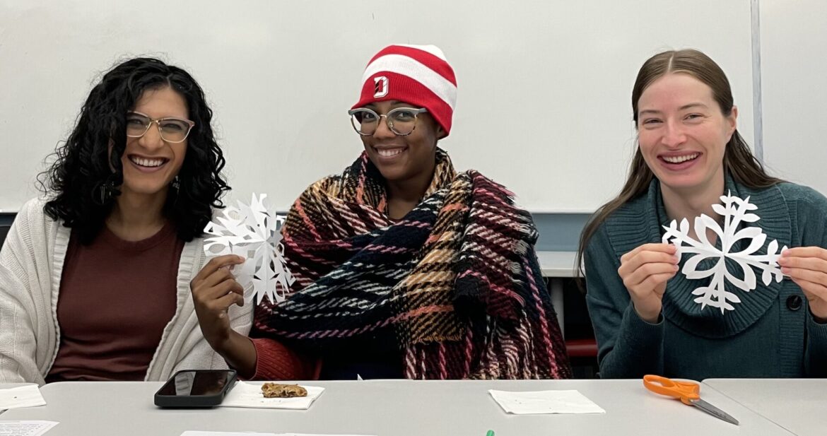 Image of three smiling friends sitting at table with paper snowflakes.