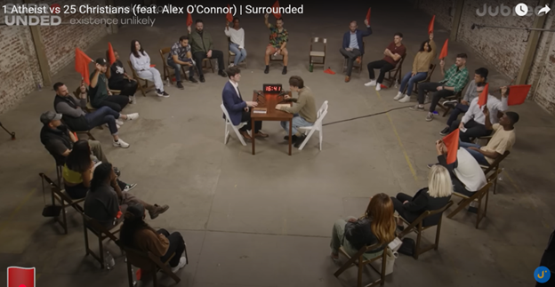 Screenshot from Surrounded, with people sitting in a circle around a table with one person at it. Some are holding red flags.