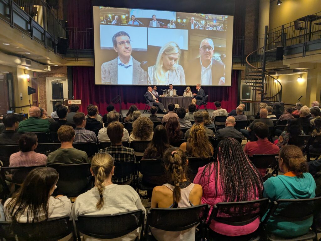 Image of roundtable with audience and screen in background
