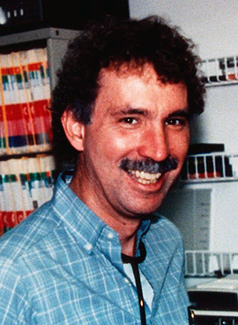 Photograph of a white male with dark hair and a mustache, wearing a blue shirt, in a science-lab setting.