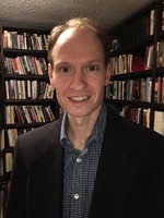 A photograph of a white male in a suit jacket, with shelves of books in the background.