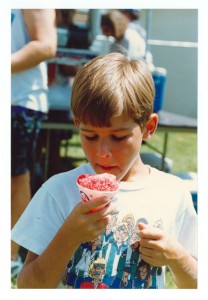 A child with light brown hair eating a Sno-Cone with some flavor of red syrup