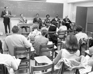 Hight teaching class, 6 professors at the front of the room, 5 sitting and 1 at a podium, while the students attentively listen