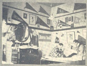 1910 dorm room, pennants and pictures on the walls. Two man sitting on the bed. There is a dresser with a large eliptical mirror on top of it. 