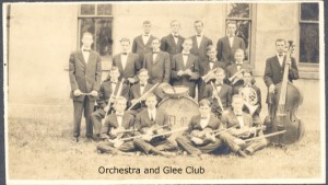 Orchestra and Glee Club, 19 men in black suits and bowties holding their instruments, violin, cello, trumpet...