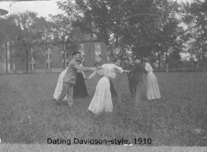 A group of people holding hands in a circle on some grass with a building and a tree in the background captioned, "Dating - Davidson style, 1910"