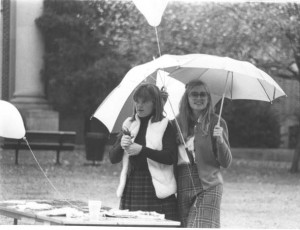 Two students holding an umbrella in one hand and a balloon in the other