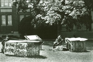 A man sitting on a chair under a tree next to a some small wooden boxes covered with graffiti that says, "No More Hypocrisy"