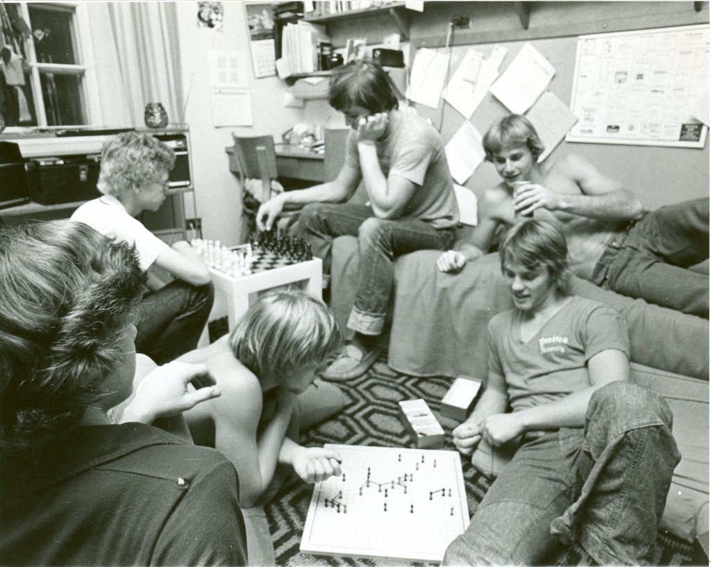 A group of students play games (chess and another unidentified game) in a Richardson dorm room in 1975 - a peek at the walls in the background reveals some typical dorm decorations, including a wall calendar.