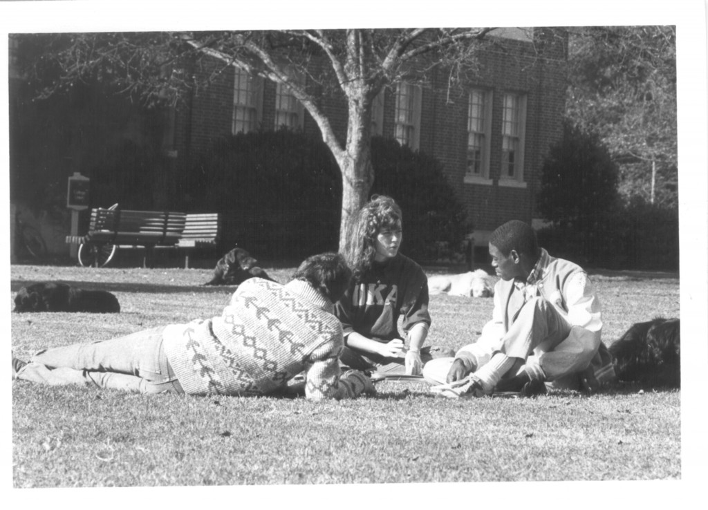 Reggit Leggett, Andrea Ward, and John Hain sit on the lawn in the grass studying and talking. There are three black dogs and one white one lounging around as well.