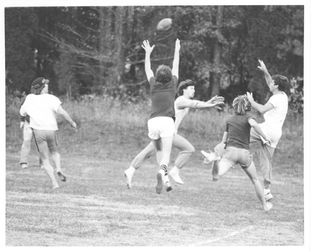 A group of students play flickerball, circa 1980s. A player has just thrown the ball to a likely reciever as he is getting downed by two defenders, the ball is still in the air while someone reaches up to catch it.