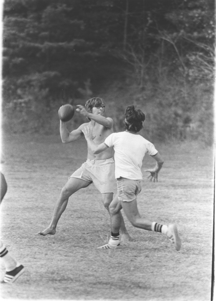 Two members of the Sigma Phi Epsilon team play flickerball, fall 1974. A shirtless man holding the football with his cocked arm ready to throw it while a defender is running toward him to stop him(he is wearing a shirt)