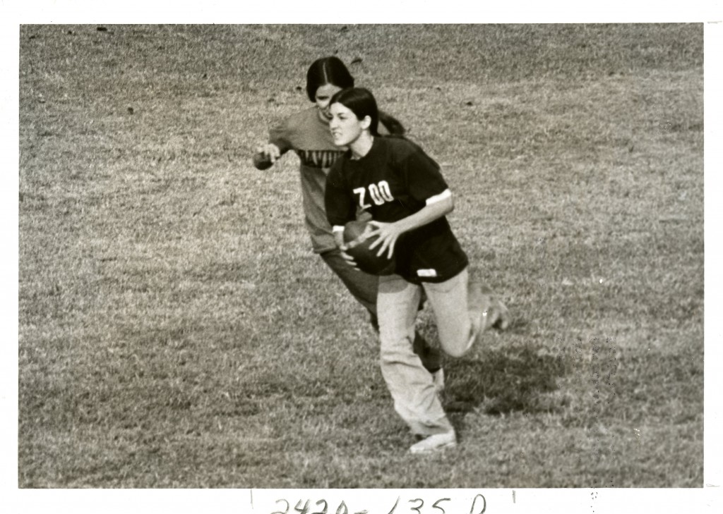 Fall 1974, the caption on this photo reads: "Girl's Flickerball - Zoo vs. Thesty." A woman with a shirt on that says, "Zoo", is holding the football running away from another player