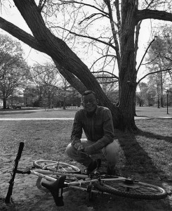 Anthony Foxx, class of 1993 squatting next to a bike on its side with a tree behind him