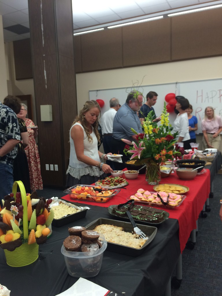 The "Luscious Brownies," amongst other goodies on a table for a retirement party. People are talking and holding plates with goodies from the table.