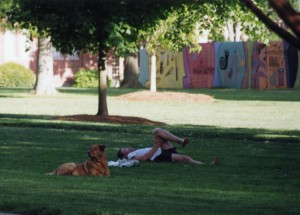 John Syme and Oscar(a dog) laying on th grass in front of campus in 2002