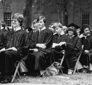 Students in the gowns(only one picutred has their cap on) sitting in chairs on the lawn waiting to graduate