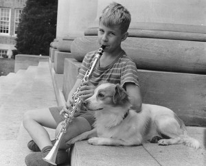 A boy playing a clarinet on the steps of chambers next to a spaniel