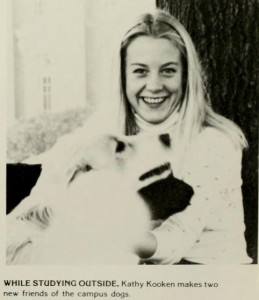 An image from a newspaper of a woman sitting next to a tree with a white dog and a black dog with the caption, "While Studying Outside, Kathy Kooken makes two new friends of the campus dogs"