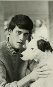 A man with a sweater and a flannel on underneath sitting next to a white dog with a brown spot on the left side of his/her head