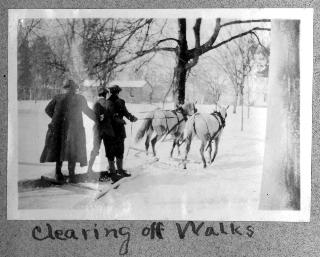 Three students clear walkways on rails pulled by horses captioned, "Clearing off Walks", circa 1915.