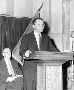 Sam Spencer as Dean of Students standing at a podium with President John Cunningham in the background.