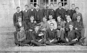 Three rows of young men in suits stand in front of windows. 