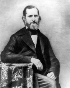 A bearded gentleman in a suit sits with his left arm folded on the armrest.