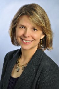 Portrait of a white business woman with short blonde hair in a gray blazer and purple blouse against a light blue background.