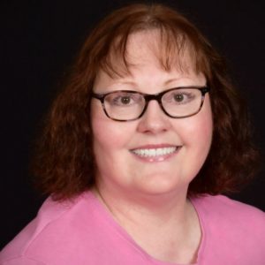 Portrait of Gillian Gremmels. Woman with black glasses, wavy brown hair with bangs and a pink blouse against a black background.