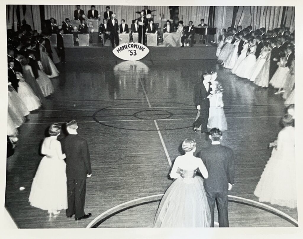 Dates on the dance floor of Homecoming 1953. At the center of the floor are three couples, all dressed in formal attire. At the edges of the floor are more couples, facing a stage where a big band is performing. 