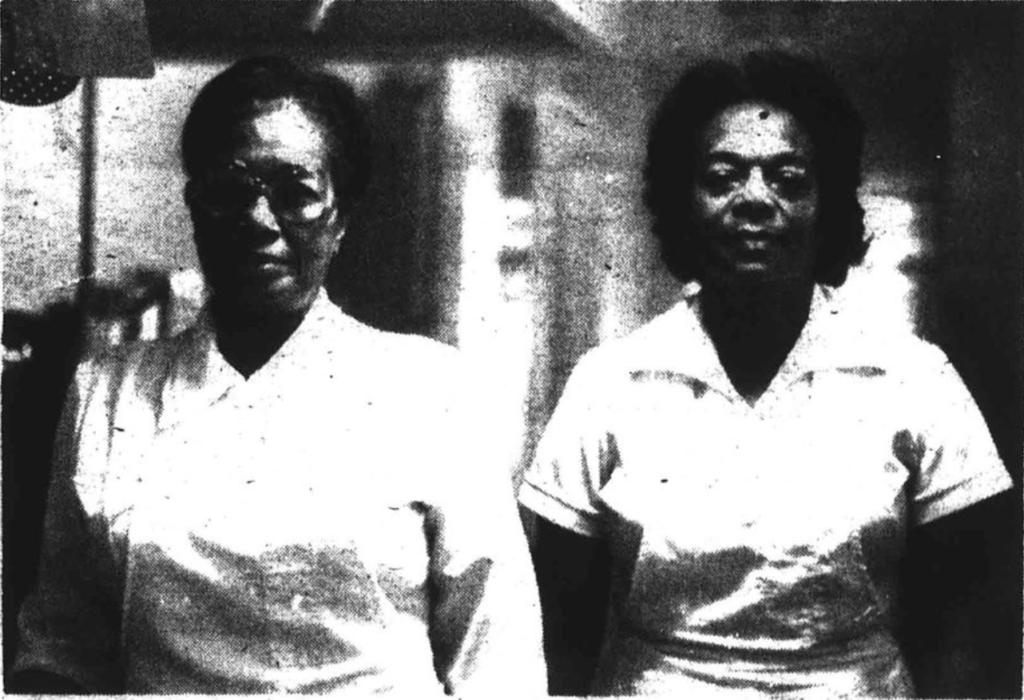 Black and white image of Fannie Brandon and Mabel Torrence. The two women are standing side by side and both are wearing white dresses with white aprons. Fannie Brandon has her hair in a bun and is wear glasses, and she is standing on the left, wearing glasses. Mabel Torrence has short hair and is standing on the right. 