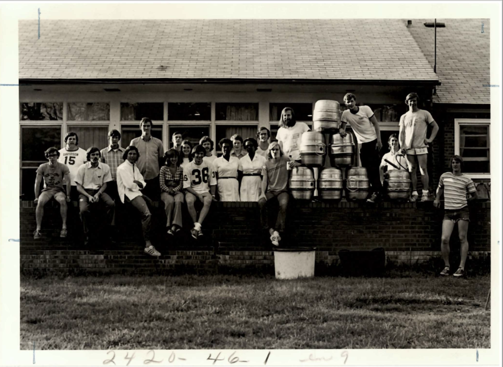 The student members of the Fannie and Mabel eating house are sitting on a brick wall outside. There are 21 people lined up, some sitting on the wall and some standing behind. There is a tower of large steel beer kegs arranged on the wall as well. Fannie and Mabel are at the center of the group. They are wearing white dresses with white aprons over them. 