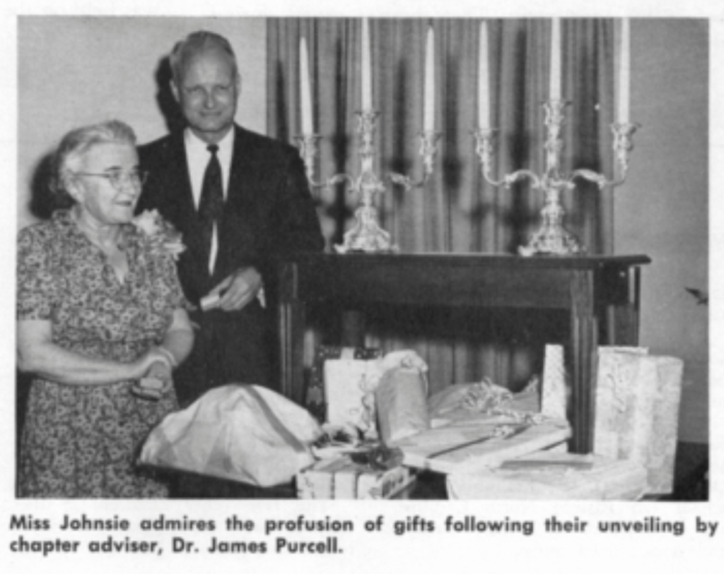 Johnsie Shelton stands next to the chapter advisor of the fraternity to accept gifts on a table in front of her. 