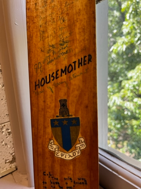 Close up on a wooden fraternity paddle. In Sharpie reads: Housemother. Underneath, in pen is the signature of J. Carey Stewart. Beneath this is the crest of the fraternity, Alpha Tau Omega. 